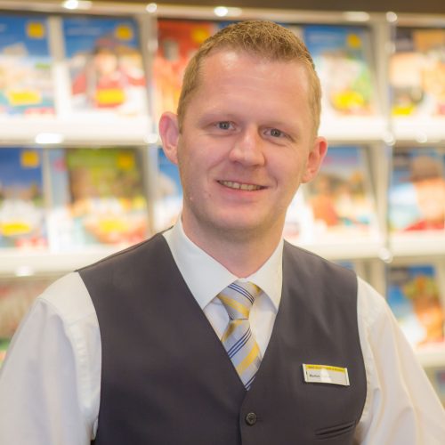 Mann in einem Anzug mit Krawatte, der in einem Geschäft lächelt und vor einem Regal mit Broschüren steht.