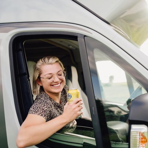 Frau mit Brille hält eine Getränkedose und lächelt aus dem Fenster eines Wohnmobils.