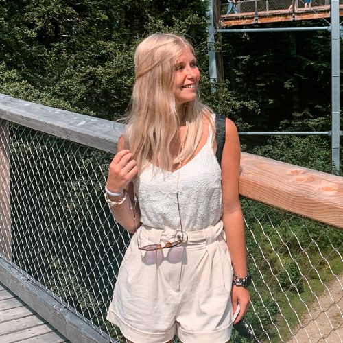 Junge Frau mit langen blonden Haaren, die auf einer Holzbrücke steht und in die Kamera lächelt.