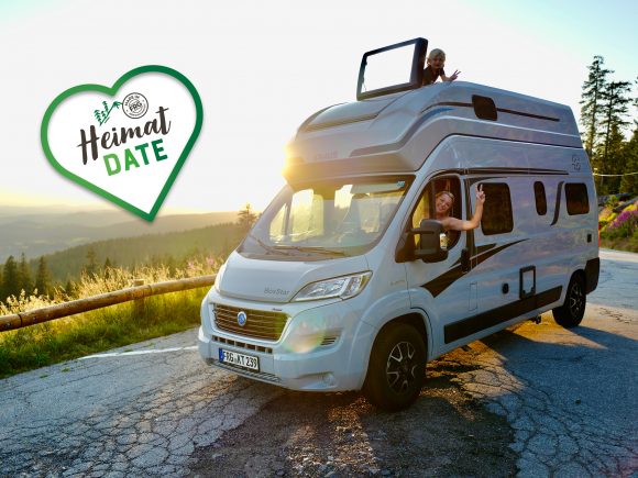 Silbernes Wohnmobil mit zwei Personen, die aus dem Fenster und dem Dach schauen, bei Sonnenuntergang.