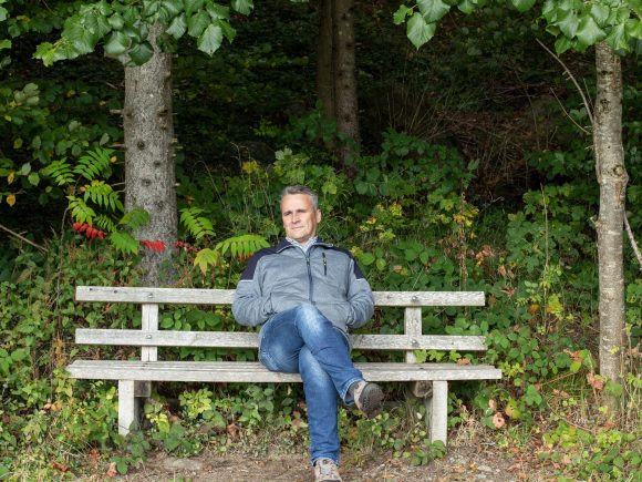 Mann sitzt entspannt auf einer Holzbank in einer grünen Umgebung mit Bäumen und Sträuchern.
