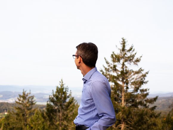 Mann mit Brille und Hemd, der in die Ferne schaut, umgeben von Bäumen und Landschaft.