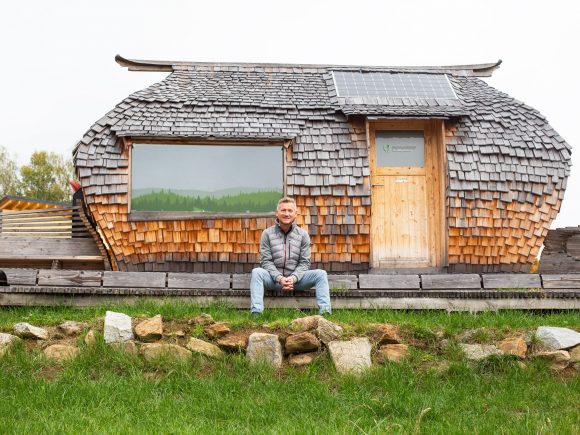 Eine Holzhütte mit einem Schindeldach und Solarzellen, davor sitzt ein Mann auf einem Stein. Die Hütte hat große Fenster.