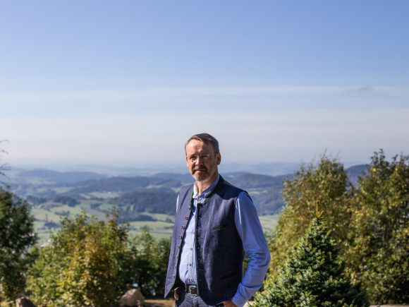 Ein Mann in traditioneller Tracht steht vor einer Berglandschaft mit Bäumen und Hügeln im Hintergrund.