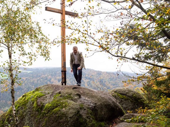 Ein Mann steht auf einem großen Felsen neben einem Holzkreuz in einer bewaldeten Umgebung.