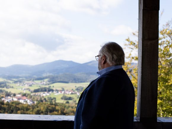 Ein Mann mit Brille steht an einem Aussichtspunkt und schaut auf die umliegende Landschaft mit Hügeln und Dörfern.