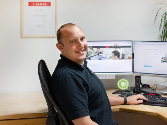 Mann sitzt an einem Schreibtisch mit zwei Computermonitoren und lächelt in die Kamera.