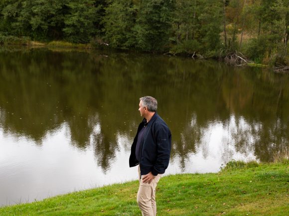 Mann steht am Ufer eines ruhigen Sees und blickt auf die Wasseroberfläche.