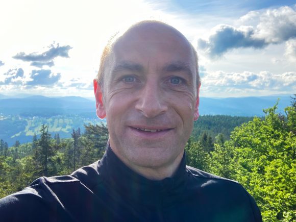 Ein Mann mit Glatze lächelt in die Kamera, während er im Freien steht, umgeben von Bäumen und Bergen.
