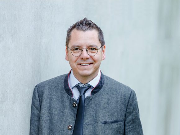 Mann mit Brille und grauem Anzug, der an einer Wand steht und lächelt.