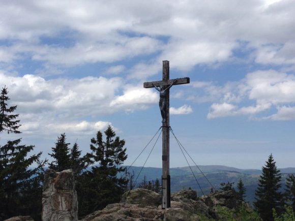 Holzkreuz mit einer Figur von Jesus Christus, auf einem Berg mit Bäumen im Hintergrund.