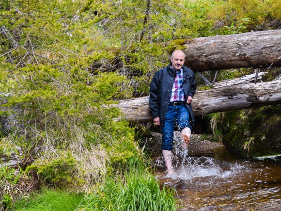 Mann sitzt am Ufer eines Baches und hat ein Bein im Wasser, während er an einem Baumstamm lehnt.