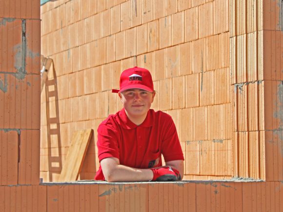 Junger Mann in rotem T-Shirt und rotem Hut, der an einer Baustelle steht und durch einen Maueröffnung schaut.