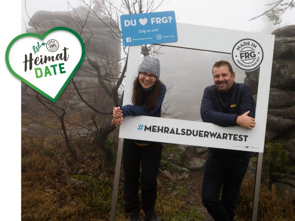 Zwei Personen posieren in einem Rahmen mit dem Text '#MEHRALSDUERWARTEST' im Freien.