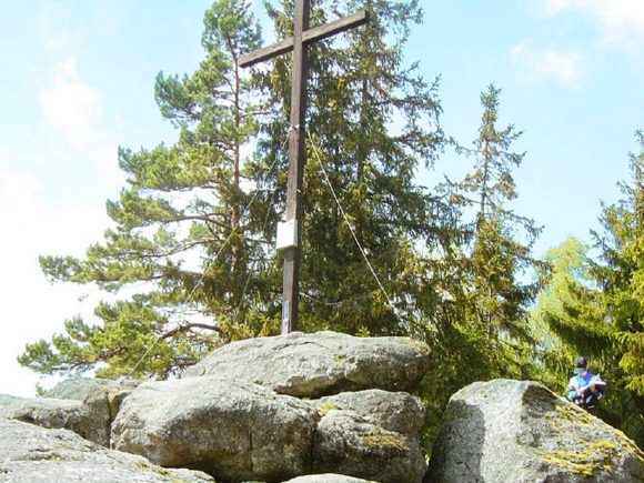 Holzkreuz auf einem großen Felsen, umgeben von Bäumen in einem Waldgebiet.