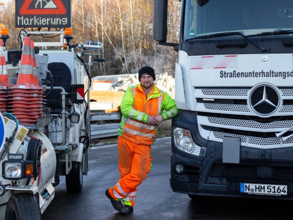 Mann in orangefarbener Arbeitskleidung steht neben einem Mercedes Lkw im Straßenbau.