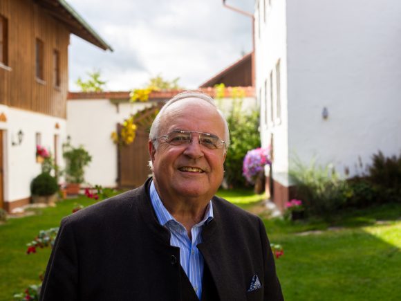 Älterer Mann mit Brille und Anzug steht lächelnd in einem Garten mit Blumen und Gebäuden im Hintergrund.