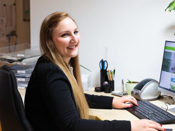 Eine Frau mit langen Haaren sitzt an einem Schreibtisch und arbeitet am Computer.