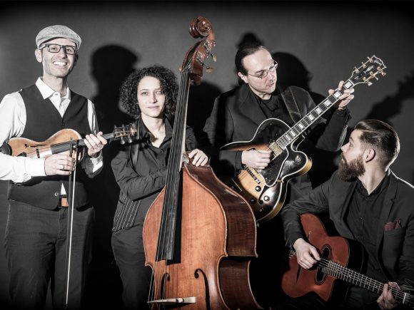 Vier Musiker mit verschiedenen Instrumenten, darunter Geige, Kontrabass, E-Gitarre und Akustikgitarre, vor schwarzem Hintergrund.