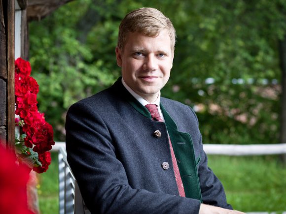 Mann in traditioneller Tracht mit einem freundlichen Lächeln und roten Blumen im Hintergrund.