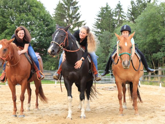 Drei Reiterinnen auf Pferden in einem Reitstall, zwei Frauen reiten braune Pferde und eine Frau reitet ein schwarzes Pferd.