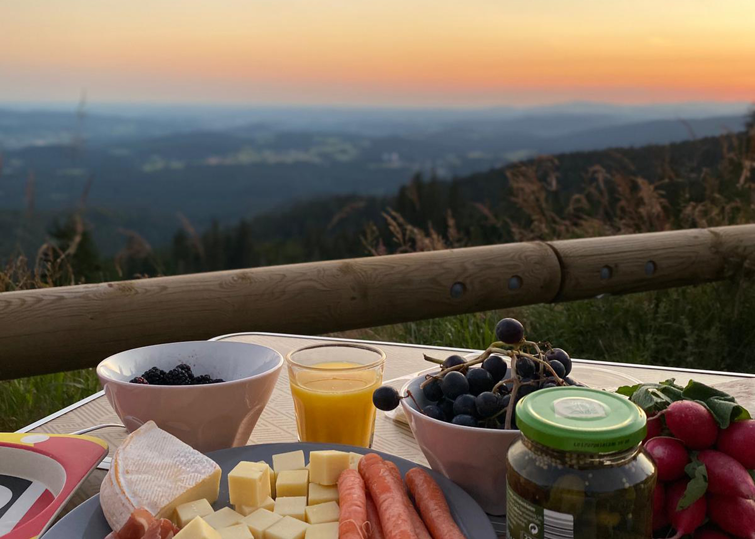 Ein Teller mit Käse, Karotten, Trauben und einem Glas Saft vor einer Landschaft bei Sonnenuntergang.