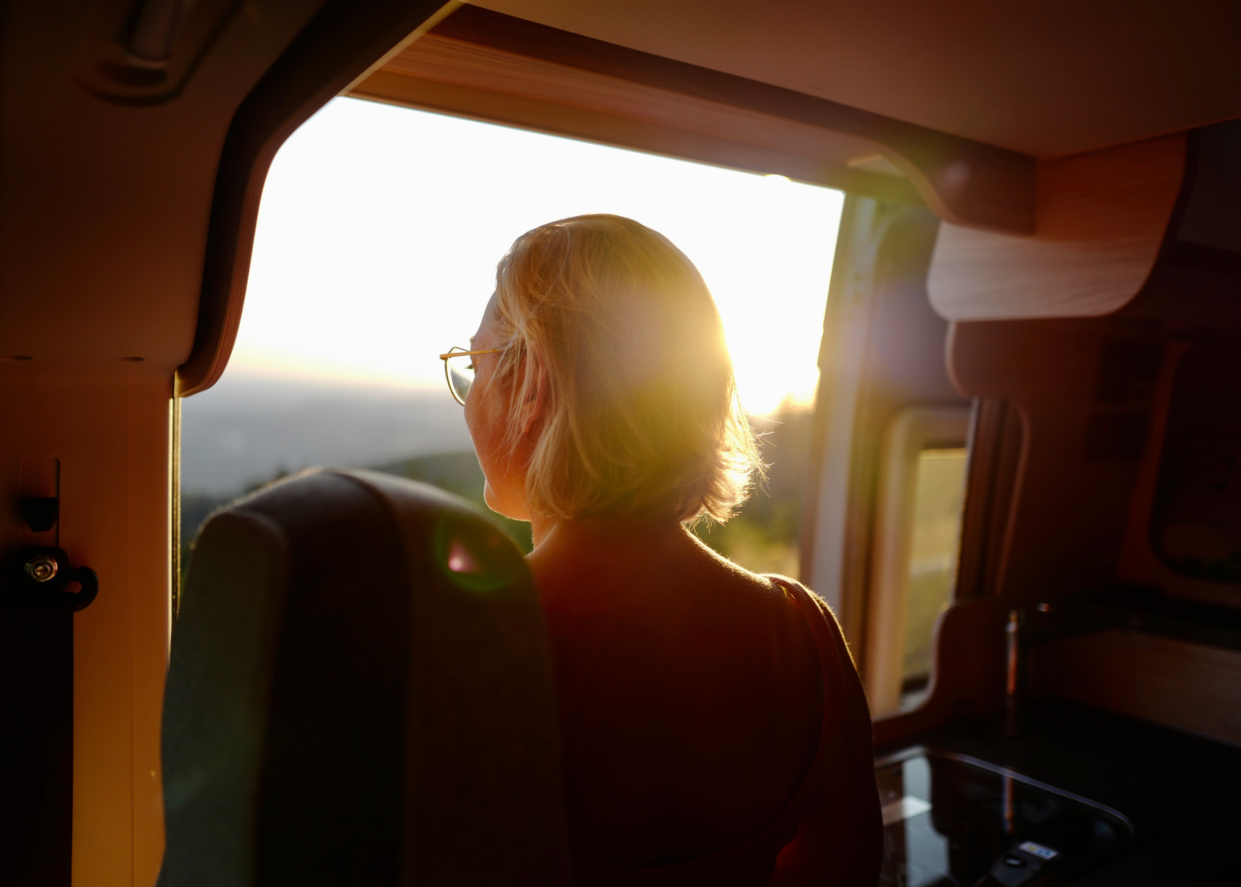 Eine Person mit Brille schaut aus dem Fenster eines Fahrzeugs während des Sonnenuntergangs.