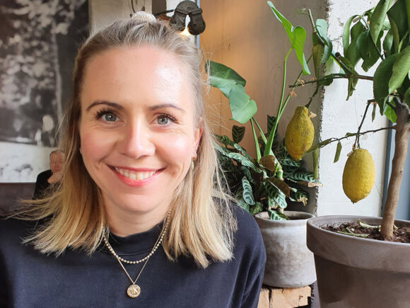 Eine Frau mit blonden Haaren und einem schwarzen Oberteil sitzt neben einem Topf mit Zitronenpflanzen.