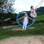 Zwei Kinder schwingen auf einer Nestschaukel in einem Spielplatzbereich mit Holzspänen.