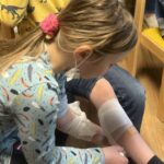 Ein Kind verbindet das Bein eines anderen Kindes mit einem Verband in einem Innenraum.