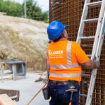 Ein Arbeiter in orangefarbener Sicherheitskleidung steht vor einer Baustelle und schaut eine Leiter hinauf.