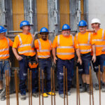 Sechs Bauarbeiter in orangefarbener Sicherheitskleidung und Helmen stehen zusammen auf einer Baustelle.