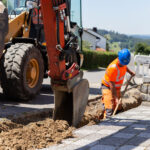 Ein Bagger und ein Arbeiter führen Straßenbauarbeiten in einem Wohngebiet durch.