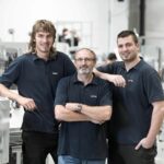 Drei Männer in blauen Poloshirts stehen in einer Fabrik mit Maschinen im Hintergrund.