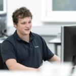 Ein Mann mit lockigem Haar sitzt an einem Schreibtisch und arbeitet an einem Computer in einem Büro.