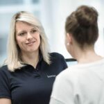 Frau mit blonden Haaren in einem blauen Poloshirt spricht mit einer anderen Person am Arbeitsplatz.