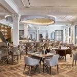 Moderne Restaurant-Innenansicht mit Bar, Tischen und Stühlen in neutralen Farben.
