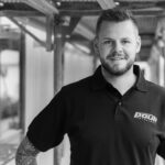 Mann mit tätowiertem Arm steht in einem schwarzen Poloshirt mit dem Logo von Pauli Metallbau.