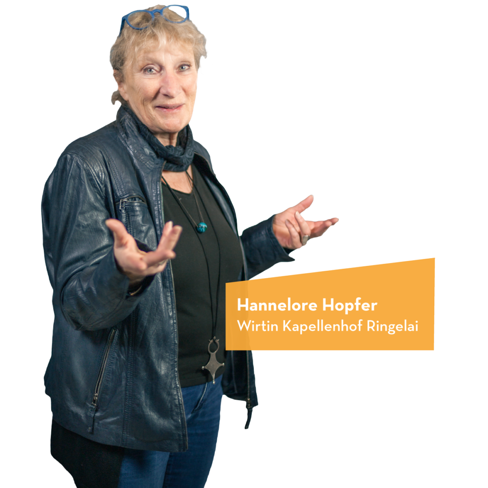 Hannelore Hopfer in einer schwarzen Lederjacke mit gestikulierenden Händen vor einem orangefarbenen Hintergrund.