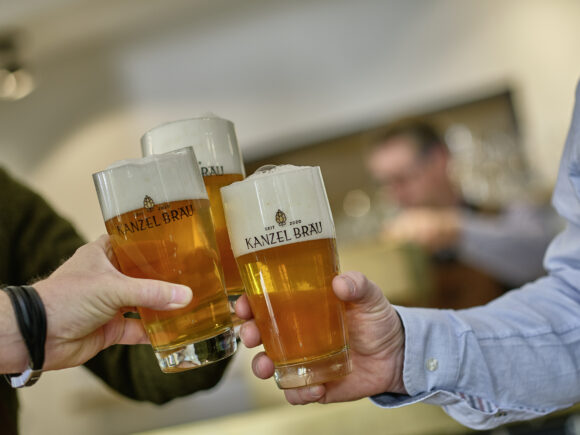 Drei Gläser mit Kanzel Bräu Bier, die zum Anstoßen gehalten werden, mit Schaumkrone.