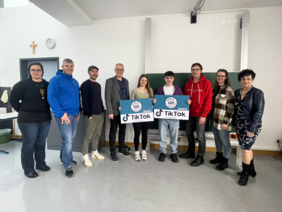 Eine Gruppe von sieben Personen steht in einem Raum und hält Schilder mit dem TikTok-Logo.