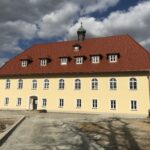 Gelbes Gebäude mit rotem Dach und mehreren Fenstern, darunter ein Eingang.