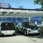 Zwei Elektroautos stehen unter einem Carport mit Solarpanelen in einer ländlichen Umgebung.