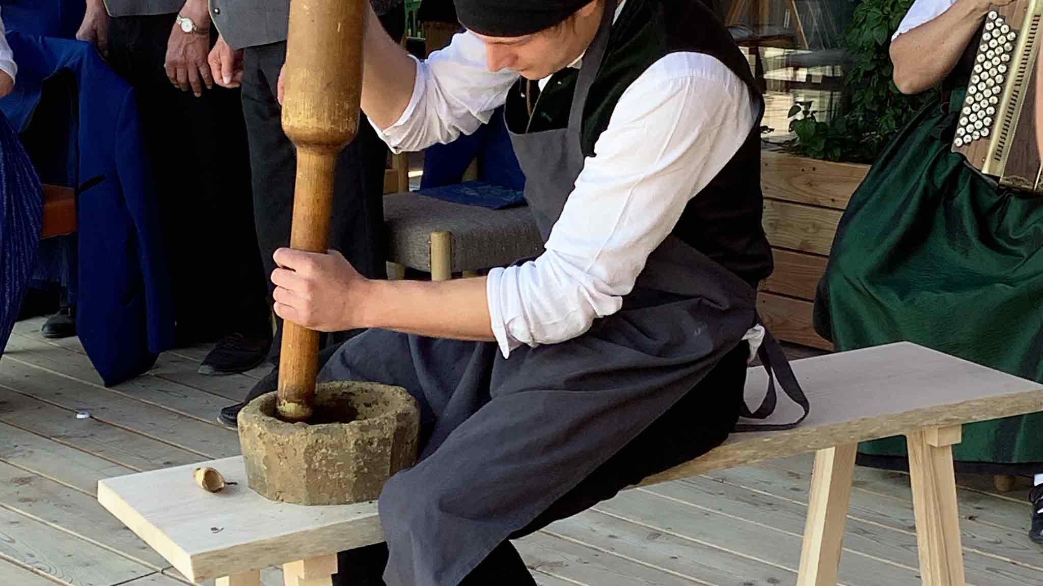 Ein Mann in traditioneller Kleidung, der einen Mörser mit einem Stößel auf einer Holzbank benutzt.