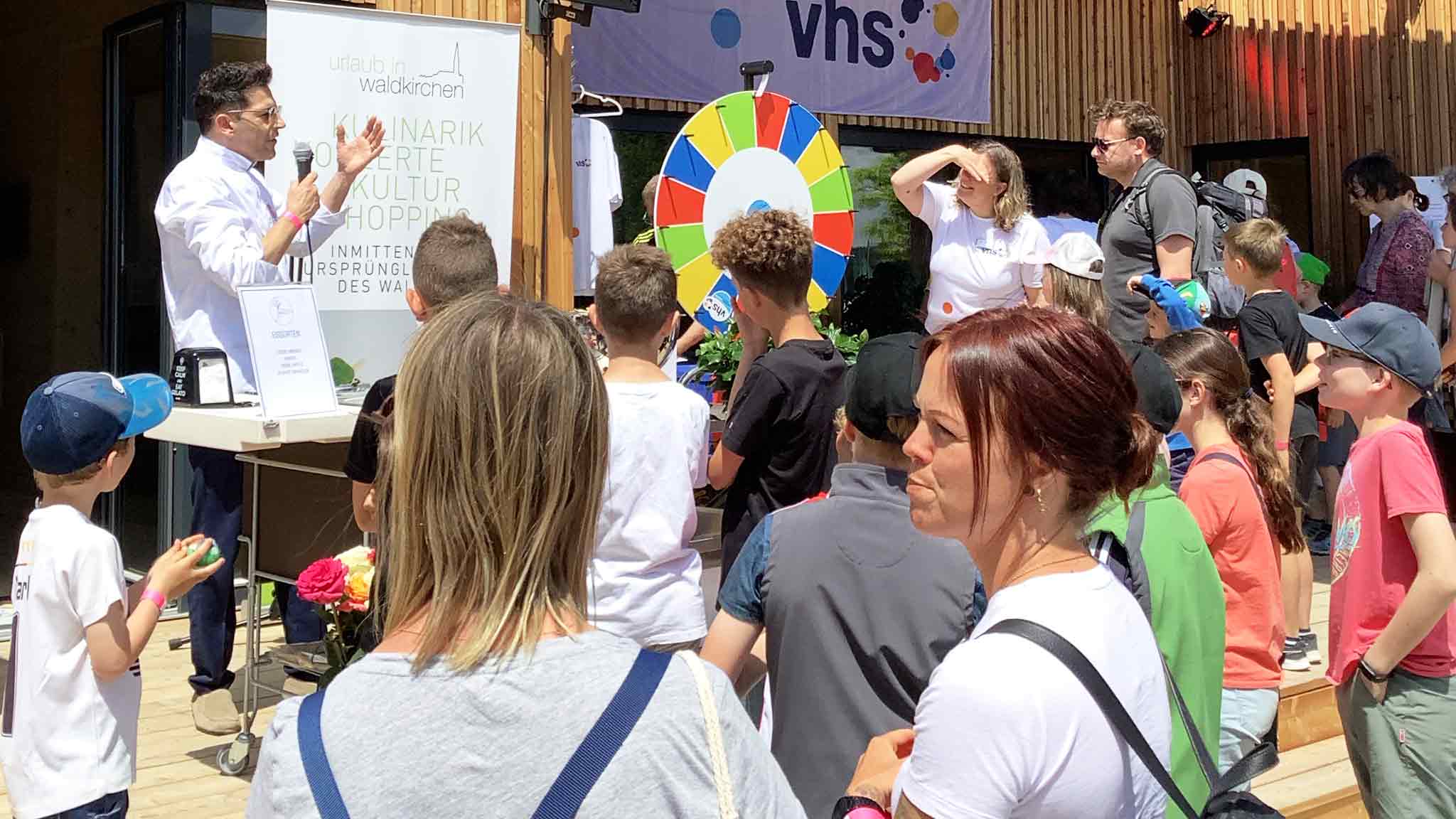 Eine Gruppe von Kindern und Erwachsenen bei einer Veranstaltung mit einem Glücksrad im Hintergrund.