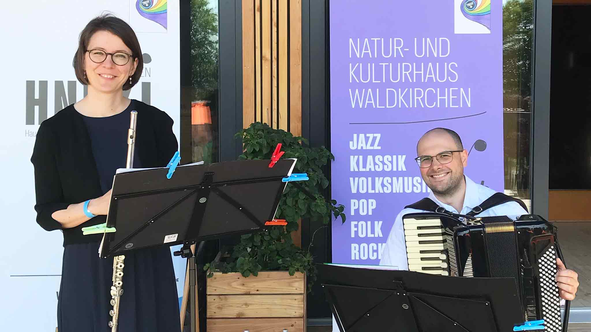 Eine Flötistin und ein Akkordeonspieler stehen vor einem Veranstaltungsort in Waldkirchen.