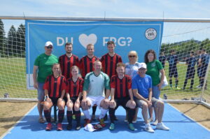 Eine Fußballmannschaft in rot-schwarzen Trikots posiert mit Fans vor einem Tor.