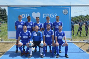Eine Fußballmannschaft in blauen Trikots posiert vor einem Tor mit einem Banner im Hintergrund.