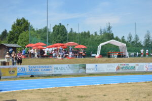 Blaue Laufbahn mit Zuschauern unter roten Sonnenschirmen bei einer Sportveranstaltung.