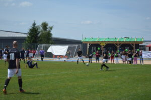 Fußballspiel mit Spielern in schwarzen Trikots auf einem Rasenfeld vor einem Tor.
